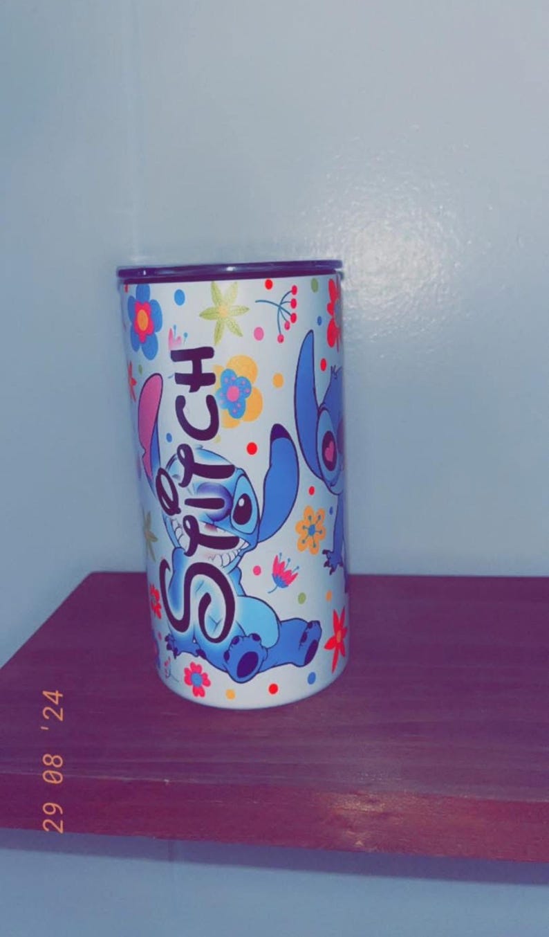 May include: A cylindrical tumbler featuring the character Stitch from the Disney movie Lilo & Stitch. The tumbler is white with a colorful floral pattern and the word "Stitch" in blue. The design includes images of Stitch.