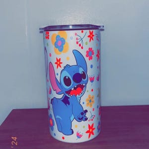 May include: A white tumbler featuring a cartoon image of Stitch from Lilo & Stitch, surrounded by colorful flowers. The blue Stitch is smiling with his mouth open. The tumbler has a silver lid.