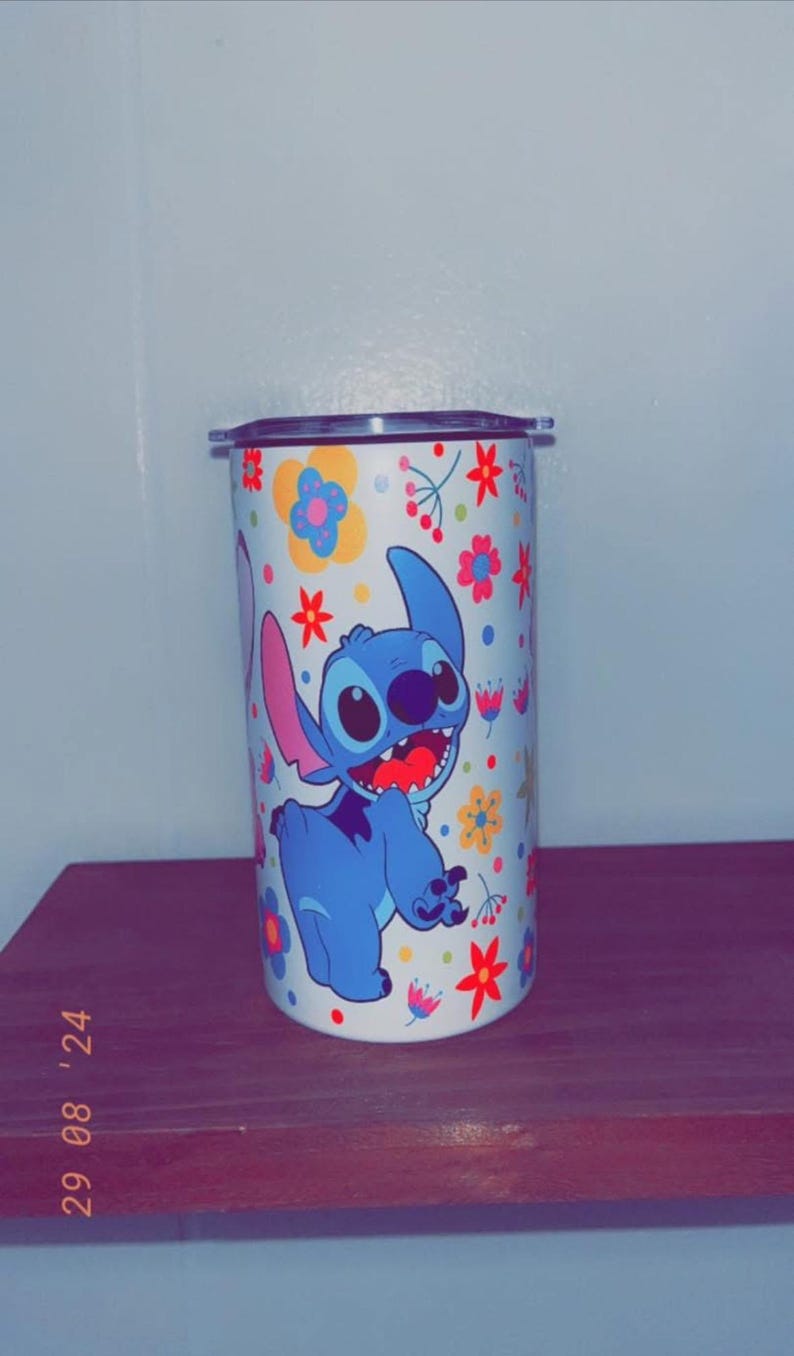 May include: A white tumbler featuring a cartoon image of Stitch from Lilo & Stitch, surrounded by colorful flowers. The blue Stitch is smiling with his mouth open. The tumbler has a silver lid.