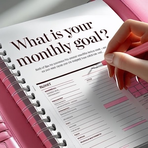 Peut inclure: Un agenda rose ouvert sur une page avec le texte « Quel est votre objectif mensuel ? » Une main tenant un stylo rose est prête à écrire sur la page. L'agenda est ouvert sur une section budget.