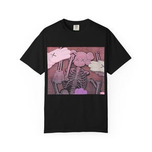 May include: Black t-shirt featuring a graphic design with a skeleton, cartoon hands, and a pink and brown background. The skeleton has crossed-out eyes. The hands are in pink, white, and gray. The shirt is a crew neck.