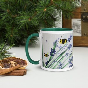 Könnte beinhalten: Weiße Keramiktasse mit grünem Rand und Henkel. Die Tasse zeigt eine Aquarellmalerei von Lavendelblüten und Bienen.