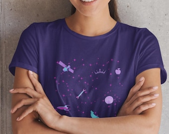 May include: A dark purple short-sleeved t-shirt featuring a graphic print of a starry night sky. The design includes small pink stars, a satellite, a crown, an apple, a wrench, a rocket ship, a moon, and a howling wolf in shades of blue and pink.
