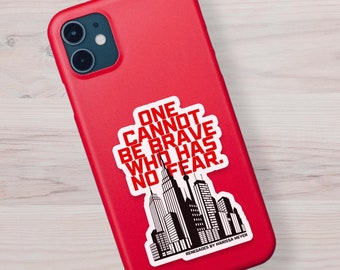 May include: Red phone case with a black and white sticker that says "ONE CANNOT BE BRAVE WHO HAS NO FEAR." The sticker also features a cityscape silhouette.
