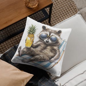 May include: A white throw pillow with a watercolor illustration of a raccoon wearing sunglasses and holding a pineapple juice. The raccoon is lounging on a beach chair.