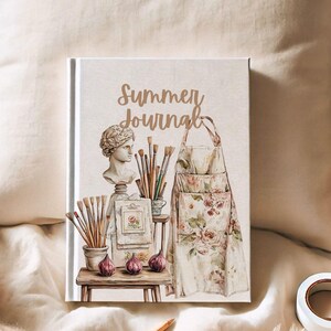 Puede incluir: Un diario de tapa dura con las palabras "Summer Journal" en escritura dorada. La portada presenta una ilustraci&oacute;n de acuarela de un delantal de artista, pinceles, un busto y otros &uacute;tiles de arte. El diario est&aacute; sobre una superficie color crema.