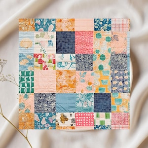 May include: A square patchwork quilt featuring a variety of patterned fabrics in shades of orange, blue, pink, and green. The quilt is composed of numerous squares, each with a unique design, including floral, seashell, and abstract patterns. The quilt is set against a white background.