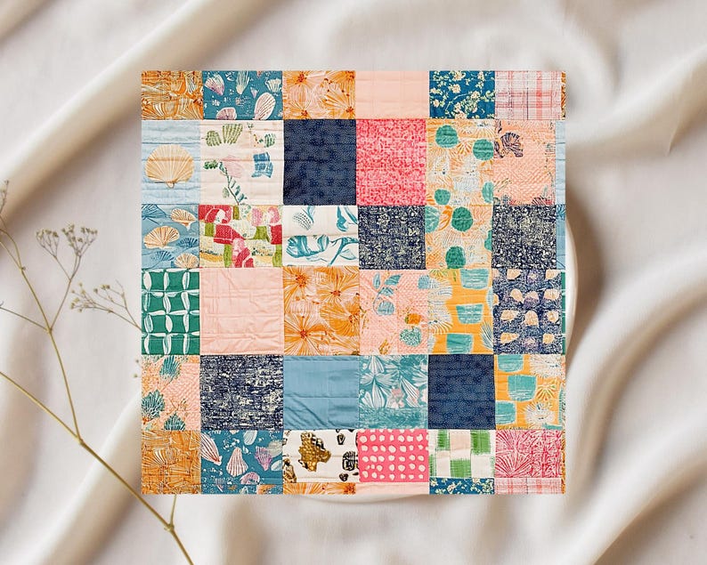 May include: A square patchwork quilt featuring a variety of patterned fabrics in shades of orange, blue, pink, and green. The quilt is composed of numerous squares, each with a unique design, including floral, seashell, and abstract patterns. The quilt is set against a white background.