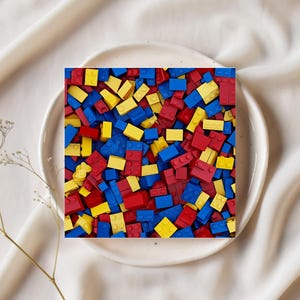 May include: A square image of a white plate filled with a colorful assortment of red, yellow, and blue toy building blocks. The blocks are scattered, creating a vibrant and playful scene. The plate sits on a white, textured surface.