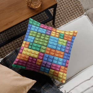 May include: Square throw pillow with a colorful pattern of interlocking toy blocks in shades of blue, green, yellow, orange, and pink. The pillow is resting on a neutral-toned surface.