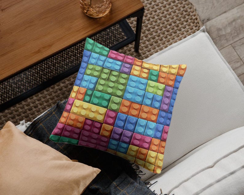 May include: Square throw pillow with a colorful pattern of interlocking toy blocks in shades of blue, green, yellow, orange, and pink. The pillow is resting on a neutral-toned surface.