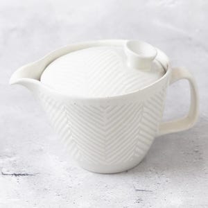 May include: A white ceramic teapot featuring a textured herringbone pattern across its body and lid. The teapot includes a curved spout, a side handle, and a small knob on the lid. The surface has a matte finish with subtle speckles throughout.