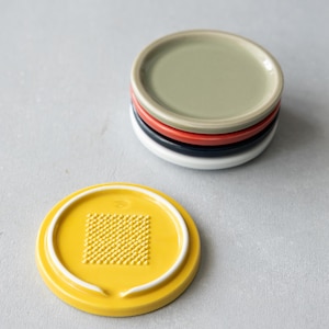 May include: A set of five circular ceramic graters in various colors including yellow, white, dark blue, red, and sage green. Each grater features a raised, textured square grid pattern in the center for grating, with a smooth, raised circular rim.