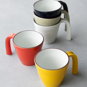 May include: A collection of five ceramic mugs with white interiors and coloured exteriors in black, beige, white, orange, and yellow. Each mug features a smooth, tapered body and a curved handle extending from the side, arranged in a group.