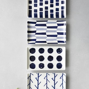 May include: A set of four rectangular ceramic plates arranged vertically. Each plate features a distinct dark blue pattern on a white background: one with vertical stripes and squares, one with geometric shapes, one with polka dots, and one with branches.
