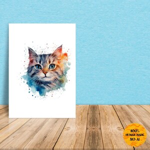 Puede incluir: Una impresión en acuarela de la cara de un gato con ojos azules y pelaje gris y naranja. La obra de arte tiene un efecto de salpicadura con pintura azul y naranja. La impresión está sobre un fondo blanco y está apoyada contra una pared azul sobre una superficie de madera.
