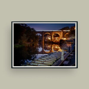 Peut inclure: Photographie encadrée d'une scène nocturne. Un pont en pierre avec des supports en arc est illuminé, se reflétant dans l'eau calme. Plusieurs bateaux sont amarrés au bord de l'eau, et le ciel passe du bleu foncé au noir.