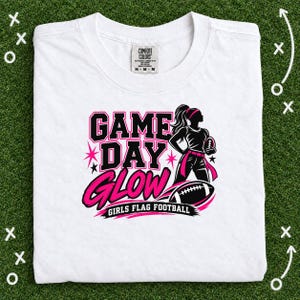 Puede incluir: Una camiseta blanca con un estampado gráfico que incluye el texto GAME DAY GLOW GIRLS FLAG FOOTBALL. El diseño presenta la silueta de una jugadora con un balón de fútbol americano, un gráfico de balón y estrellas en negro y rosa.