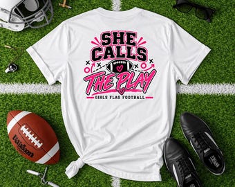 May include: A white short-sleeved t-shirt featuring a graphic design in black and pink. The text reads "SHE CALLS THE PLAY" with a football illustration and the words "GIRLS FLAG FOOTBALL" printed below in a bold, athletic font style.