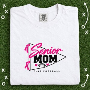 Puede incluir: Una camiseta blanca de manga corta con un gráfico de un banderín con el texto Senior MOM y FLAG FOOTBALL. El diseño incluye lazos rosas, dos pequeños corazones rosas y una ilustración de un balón de fútbol americano en tinta negra y rosa.