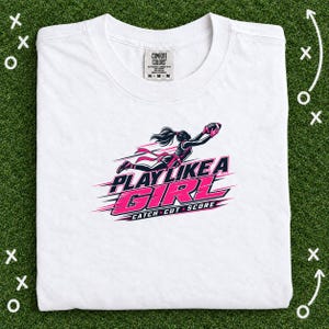 Puede incluir: Una camiseta blanca con un gráfico de una jugadora de fútbol americano en movimiento con detalles en rosa y negro. El texto dice PLAY LIKE A GIRL y CATCH CUT SCORE. La etiqueta de la marca Comfort Colors en el cuello indica talla medium.
