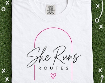 May include: A white short-sleeve t-shirt featuring a pink arched graphic with the black text She Runs ROUTES, a small pink heart, and the text PLAY. EMPOWER. BELONG. in black. The shirt has a Comfort Colors brand label at the inner neckline.