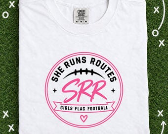 May include: A white short-sleeve t-shirt featuring a circular graphic with the text "SHE RUNS ROUTES" in black, "SRR" in large pink script, and "GIRLS FLAG FOOTBALL" in black on a pink banner, with a football icon and a small heart symbol.
