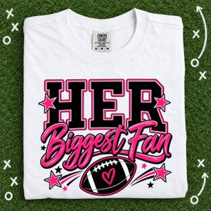 Puede incluir: Una camiseta blanca de manga corta con un diseño gráfico que incluye el texto negro HER en fuente negrita, el texto rosa en cursiva Biggest Fan y una ilustración de un balón de fútbol americano negro con un corazón rosa y estrellas.