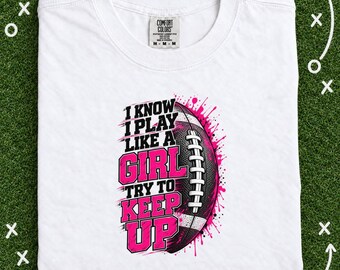 May include: A white short-sleeved t-shirt featuring a graphic of a football and the text I KNOW I PLAY LIKE A GIRL TRY TO KEEP UP in black and pink lettering. The design includes pink paint splatter accents and a Comfort Colors brand label.
