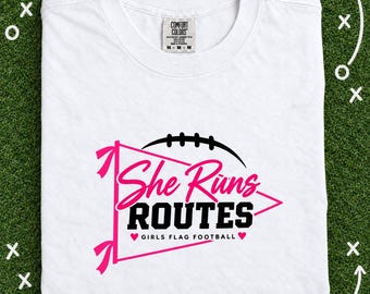 May include: A white t-shirt featuring a pink pennant graphic with a black football icon above it. The text reads "She Runs ROUTES" in pink and black, with "GIRLS FLAG FOOTBALL" and two small pink hearts printed below the text.