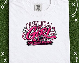 May include: A white short-sleeved t-shirt featuring a graphic design in pink, black, and white. The text reads "PLAY LIKE A GIRL" and "SHE RUNS ROUTES" with a football illustration and stylized arrows. The shirt has a Comfort Colors brand label.