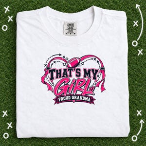 Puede incluir: Una camiseta blanca de manga corta con un estampado gráfico que incluye un balón de fútbol americano rosa, cintas y el texto "THAT'S MY GIRL PROUD GRANDMA". El diseño incluye diagramas de juego en negro y rosa y corazones, con etiqueta Comfort Colors.