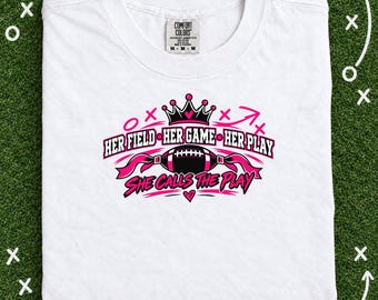 May include: A white short-sleeve t-shirt featuring a black and pink graphic design. The print includes a crown, a football, and the text: HER FIELD HER GAME HER PLAY SHE CALLS THE PLAY. The shirt has a Comfort Colors brand label at the neck.