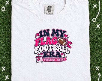May include: A white t-shirt featuring a graphic design with the text "IN MY FLAG FOOTBALL ERA" and "WEEKENDS BOOKED". The design includes a brown football, pink hearts, and a calendar icon, all rendered in a bold, retro-style font with pink and navy blue colors.