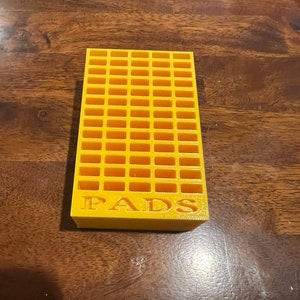 May include: A yellow plastic storage organiser with a grid pattern and the word "PADS" printed on the front.