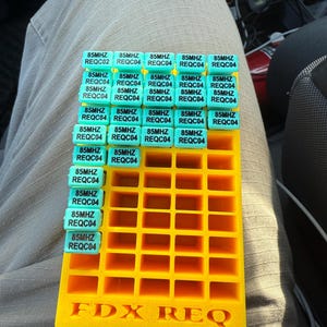 May include: A yellow plastic storage box with a grid of compartments. The compartments are filled with small, turquoise blue labels that say "85MHZ REQC04". The box has the words "FDX REQ" printed on the bottom.