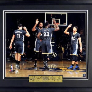 May include: A framed photograph of four basketball players in uniform, with the text "Golden State Warriors 2015 Champions" on a gold plaque at the bottom. The players are wearing black and gold uniforms with the numbers 11, 23, and 30.