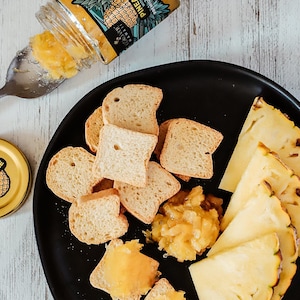 May include: A black plate with pineapple slices, toasted bread, and a jar of pineapple spread. The jar has a yellow label with the text "PINEAPPLE PREMIUM FRUIT SPREAD WITH GUAVA" and "KOLOI HAWAII MADE ON OAHU".