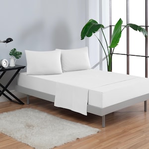 May include: A white bed with a white bedspread and two white pillows. The bed is made up with a white fitted sheet and a white flat sheet. The bed is sitting on a wooden floor. There is a plant in the background.