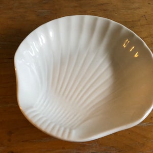 Pode incluir: Um prato branco em forma de concha com um design estriado em forma de leque. O prato de cerâmica está vazio e assenta numa superfície de madeira. O prato é provavelmente usado para servir pequenas porções de comida ou como um item decorativo.