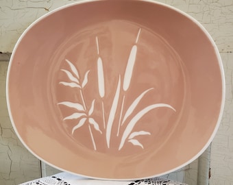 Pode incluir: Um prato redondo cor-de-rosa com uma borda branca. O prato apresenta um gráfico branco de juncos e caniços. O prato está sobre uma toalha de mesa de renda branca, com um fundo branco desgastado.
