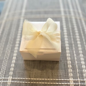May include: A white gift box with a white satin ribbon tied in a bow. The box is sitting on a gray and white patterned surface.