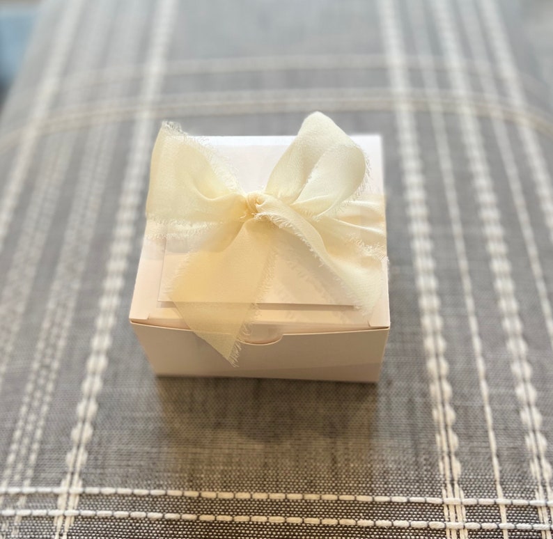 May include: A white gift box with a white satin ribbon tied in a bow. The box is sitting on a gray and white patterned surface.