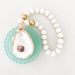 May include: A white oyster shell with a gold rim, hanging from a white and gold beaded garland. The oyster shell is on a teal blue glass base.