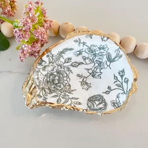 May include: A white ceramic dish with a gold rim and a black and white floral pattern. The dish is shaped like a seashell.