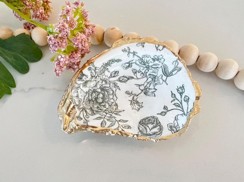 May include: A white ceramic dish with a gold rim and a black and white floral pattern. The dish is shaped like a seashell.