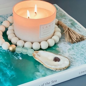 May include: A white beaded garland with a gold bead and a light blue glass bead. The garland is wrapped around a pink candle with a white label that reads "SAND", "MANGO", and "TANGERINE". The candle is on a book with a blue and green ocean wave design. The book has the text "Lewis Blackwell" and "of the Sea" on the cover. The garland has a brown tassel.