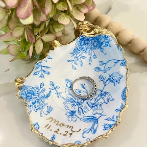May include: A white ceramic ring dish with a blue floral pattern and gold trim. The dish has the word "mom" and the date "11.2.24" written on it.