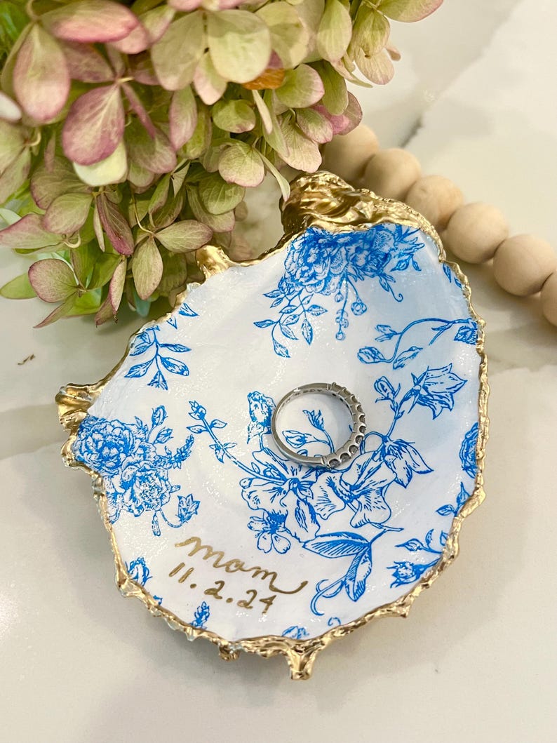 May include: A white ceramic ring dish with a blue floral pattern and gold trim. The dish has the word "mom" and the date "11.2.24" written on it.