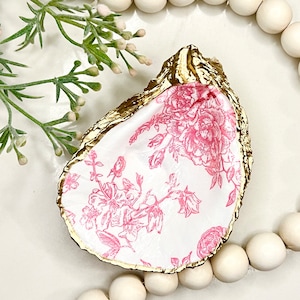 May include: A gold-rimmed oyster shell dish with a pink floral pattern. The dish is sitting on a white surface with a string of wooden beads around it.